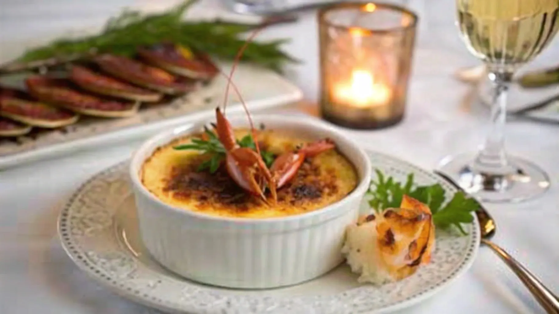 Elegant serving of crab brulee in a ramekin, garnished with fresh herbs and shrimp, accompanied by a side of roasted bread and a lit candle in the background.