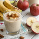 Banana and apple smoothie in a glass with cinnamon dusting and apple slice, set against a bright kitchen backdrop