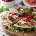 Colorful watermelon pizza topped with berries, kiwi, coconut flakes, and mint leaves on a rustic wooden board