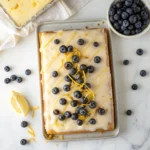 Overhead view of lemon blueberry sheet cake on a white marble background, decorated with glaze, blueberries, and lemon zest.
