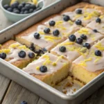 Close-up of freshly baked lemon blueberry sheet cake topped with lemon glaze, blueberries, and lemon zest in a rectangular pan.